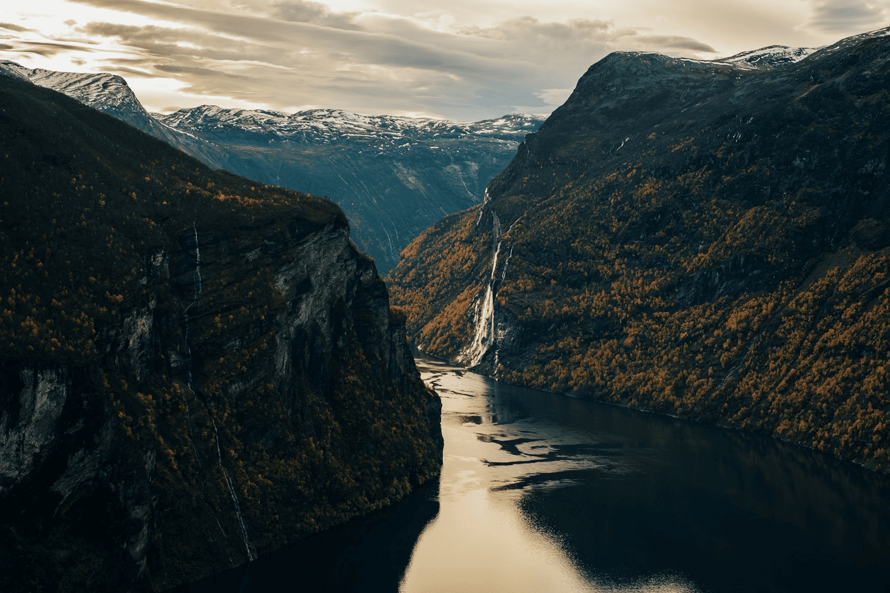A scenic view of a narrow fjord surrounded by steep cliffs and mountains under a cloudy sky.
