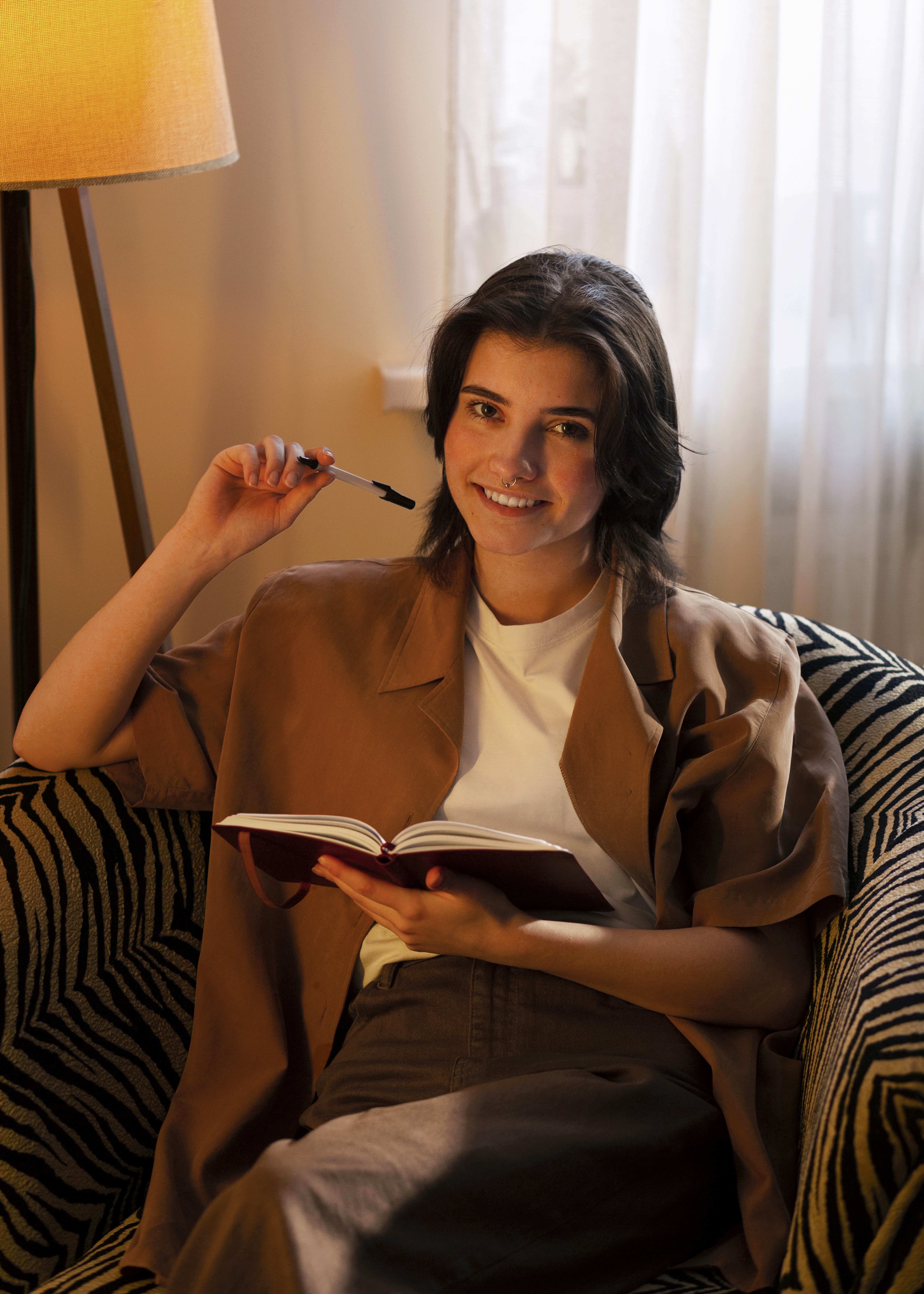 Eine Frau sitzt in einem Sessel. Sie hält Notizbuch und Stift in den Händen und lächet in die Kamera. Im Hintergrund sorgt eine Lampe für eine gemütliche Atmosphäre.