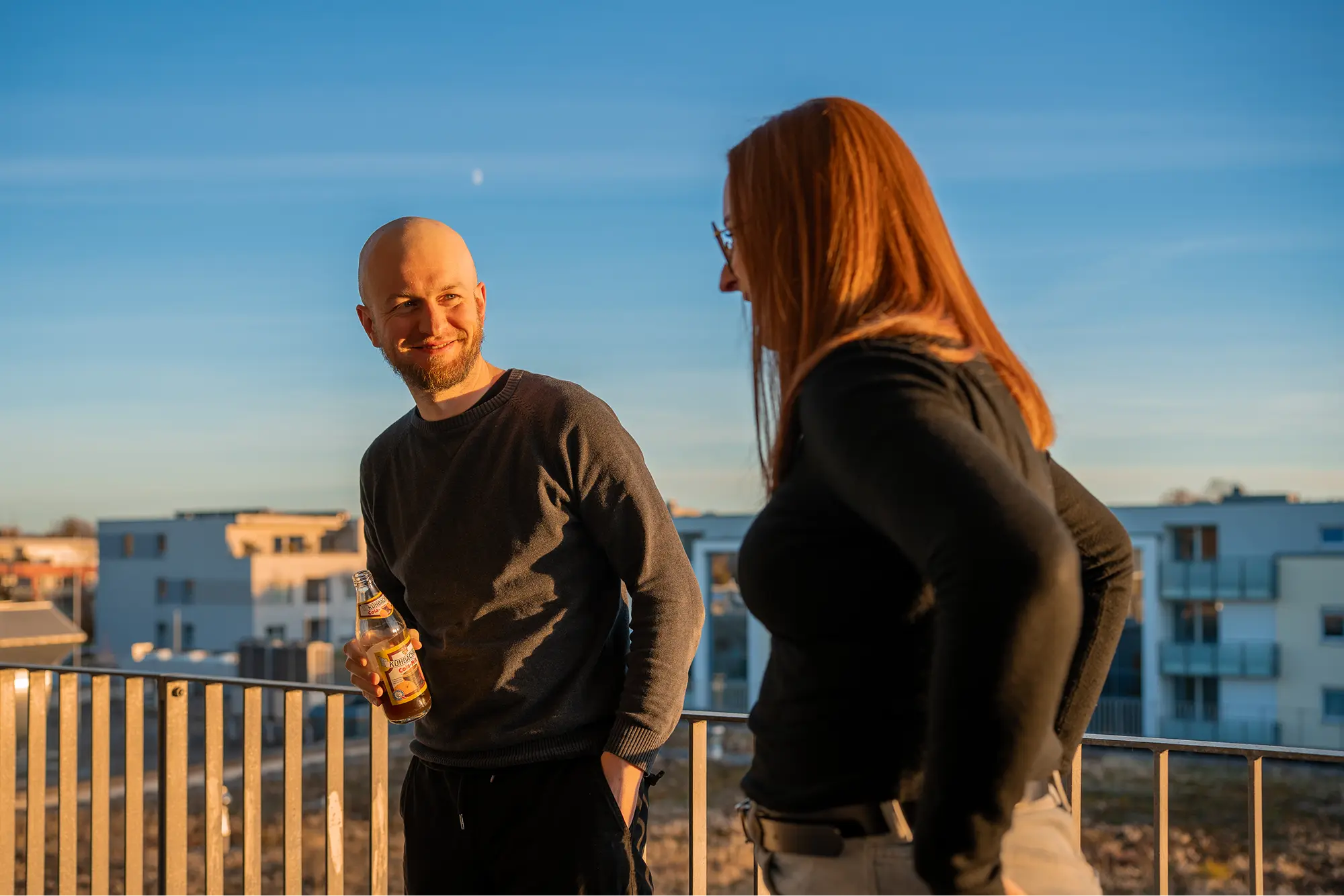 Zwei lächelnde Teammitglieder bei einem lockeren Gespräch auf einer sonnigen Dachterrasse am späten Nachmittag vor dem Panorama von städtischen Wohngebäuden.