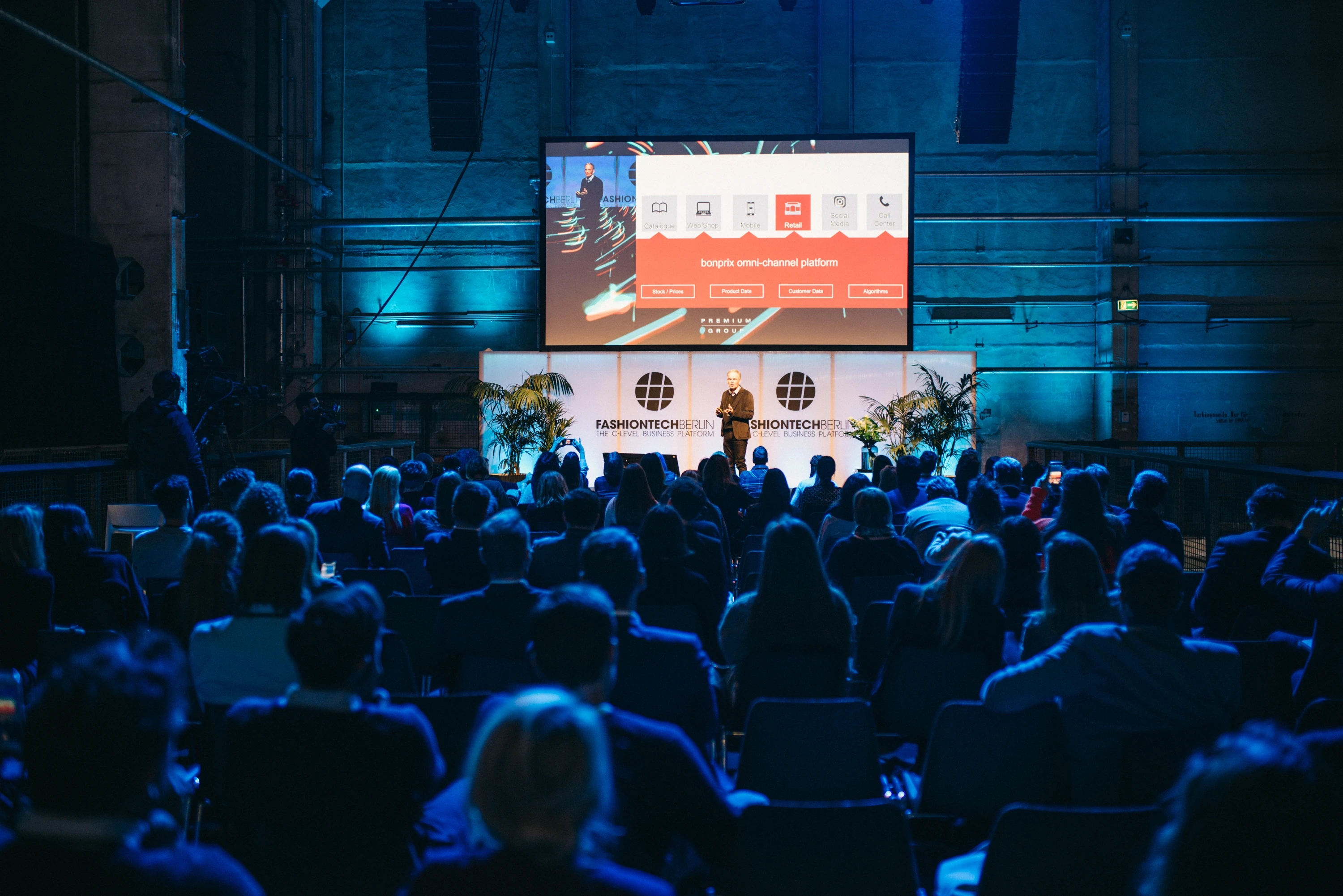 Speaker on the FASHIONTECH Berlin stage in front of a large presentation screen, with the audience in blue light in the Kraftwerk hall.