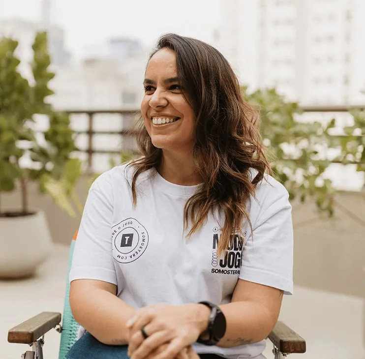 Uma pessoa sorridente com cabelos longos está sentada em uma cadeira, cercada por plantas e com uma vista da cidade ao fundo.