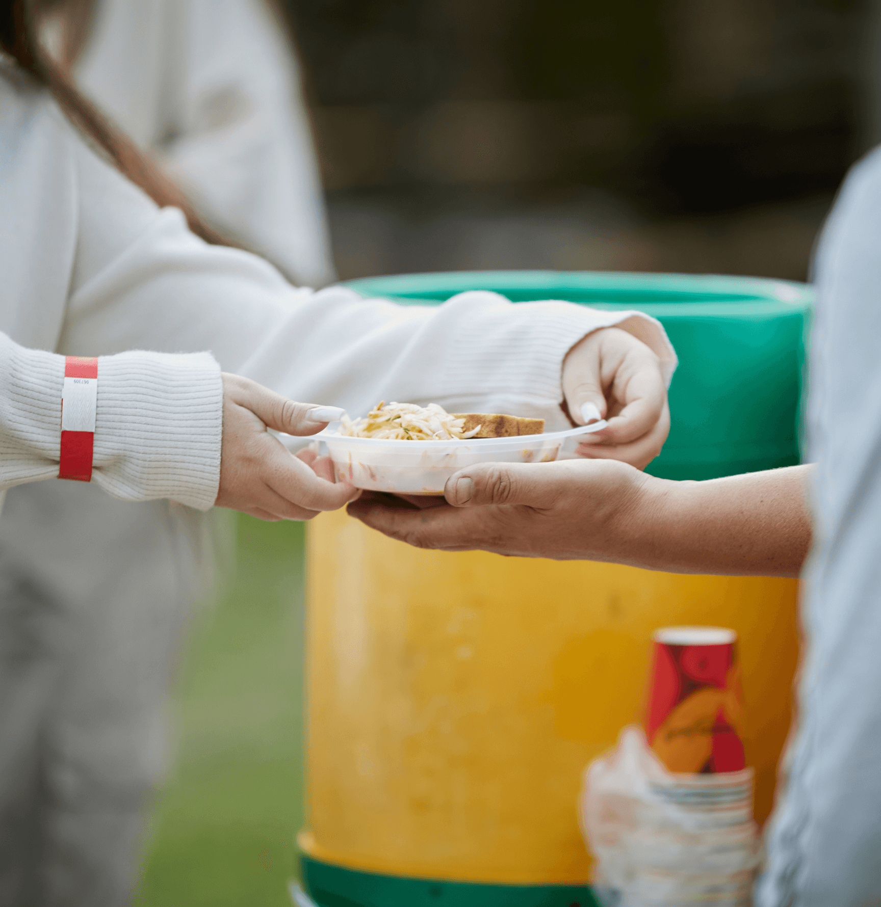 A hand is offering food on a plate to another hand near colorful barrels in an outdoor setting.