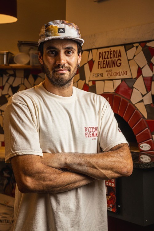 A man with crossed arms smiles while standing in a casual setting with shelves and decorations in the background.