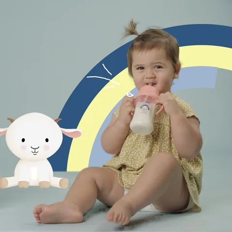 A toddler drinks from a cup, a drawn goat and rainbow beside her