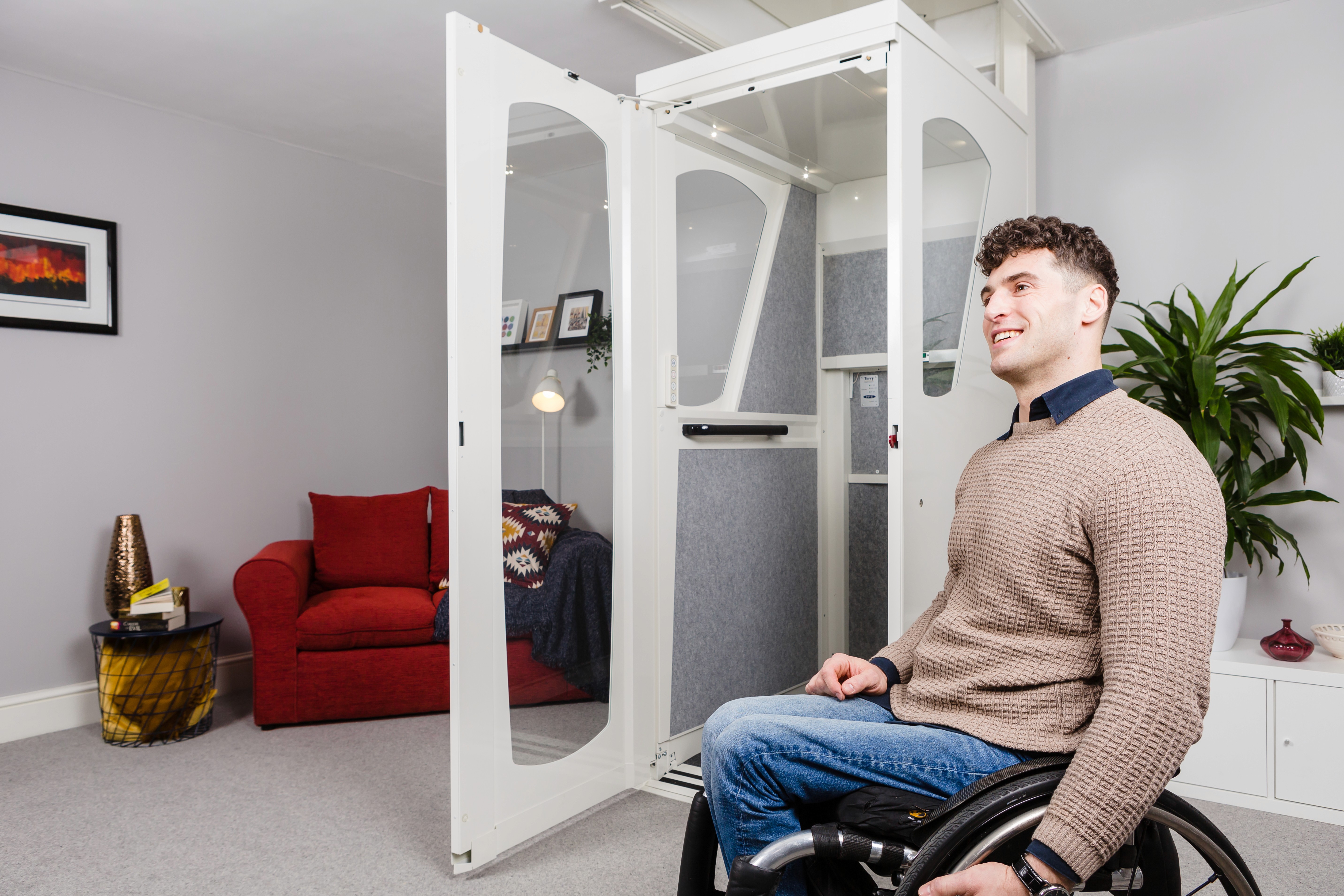 Wheelchair user smiling beside open Elevate AVA home lift in a residential living room — white through-floor lift with door fully open and interior visible