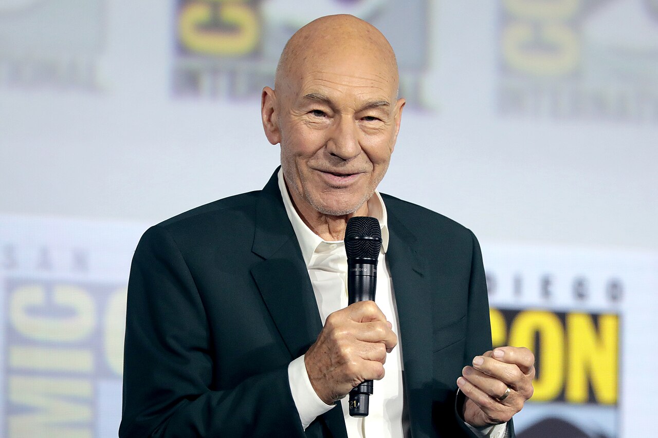 Patrick Stewart in a dark green suit holds a microphone, speaking at an event. The background features a stage with branding elements and logos.