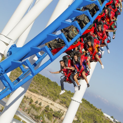 Personas montando una montaña rusa roja y azul, suspendidos boca abajo, con árboles y un cielo claro de fondo.