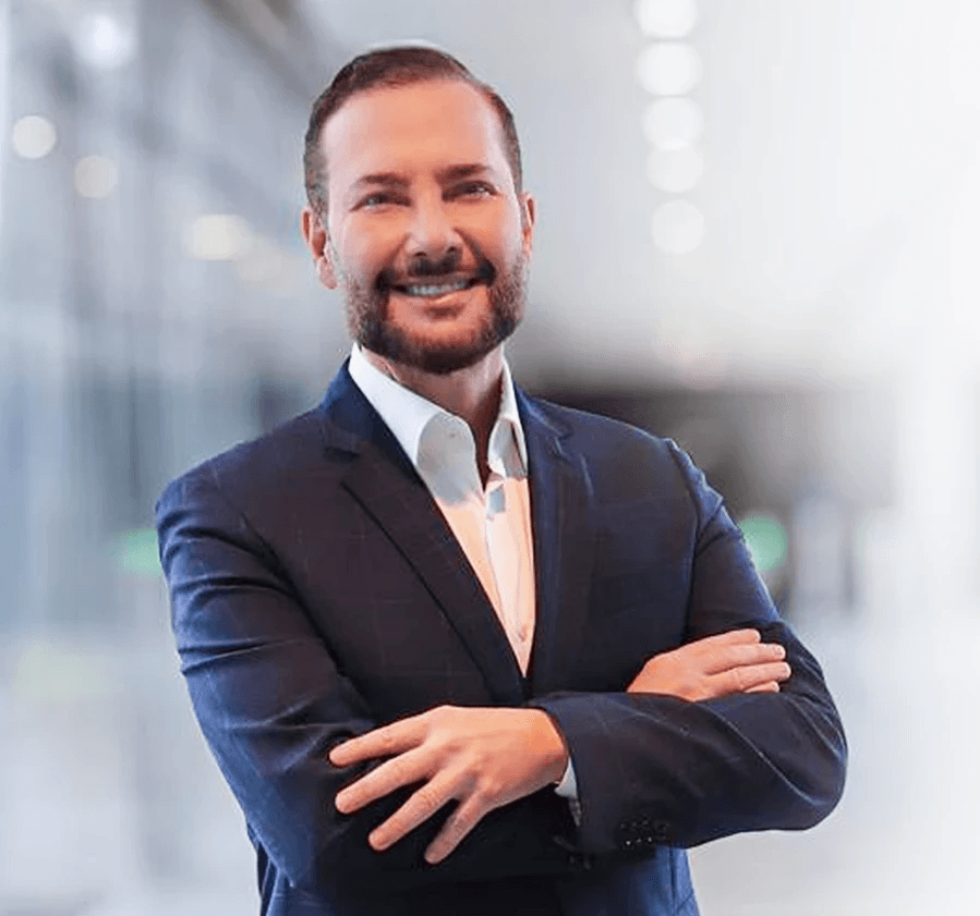 A young person with curly hair smiles at the camera while sitting in front of a laptop against a dark background.