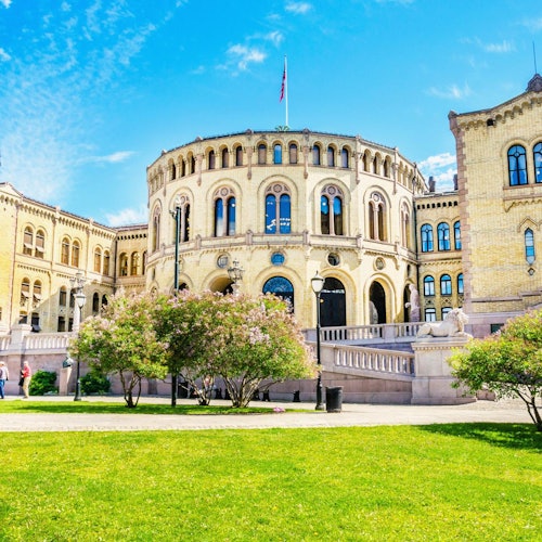 A historic stone building with arched windows and a flag on top, surrounded by green lawns and blooming trees under a clear sky.