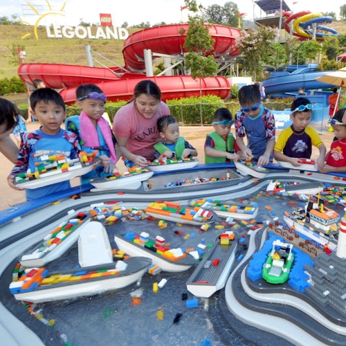 Children and an adult play with floating LEGO bricks in a water-themed play area at LEGOLAND, with large water slides in the background.