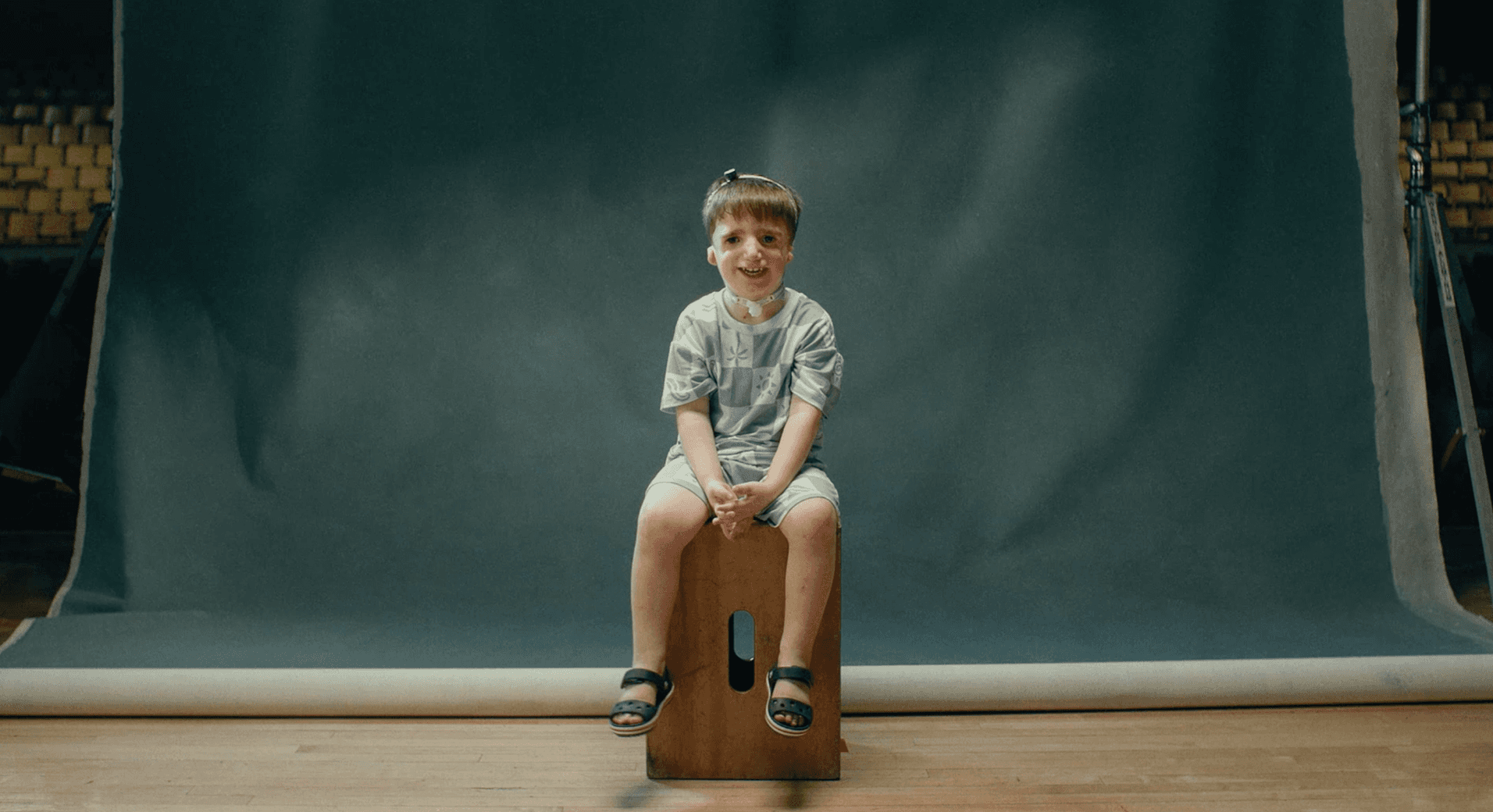 A young girl sits on a wooden crate against a blue backdrop, wearing a light-colored outfit and looking thoughtfully ahead.