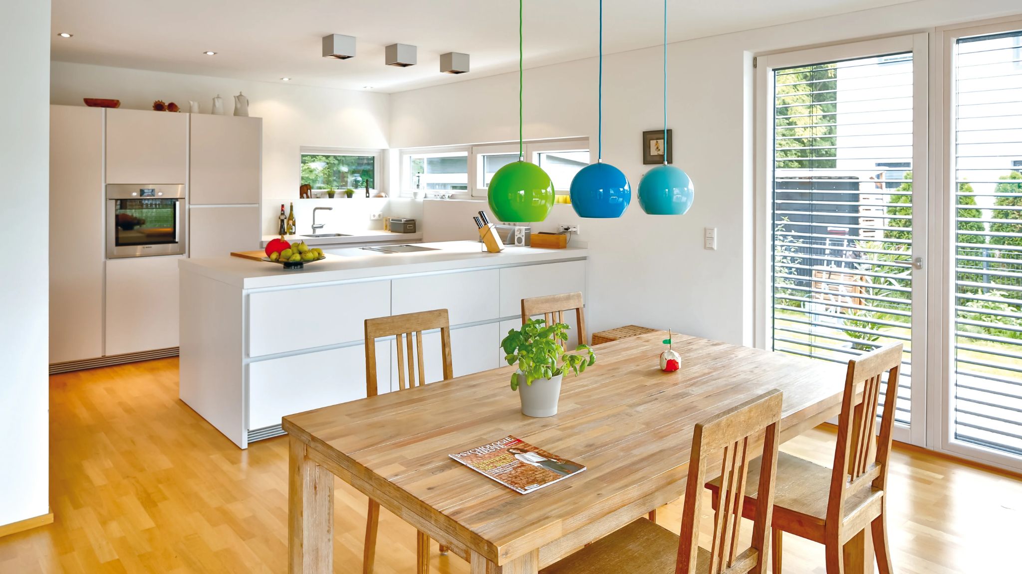 Helles und modernes Esszimmer mit Kücheninsel, Holztisch und bunten Hängeleuchten über dem Essbereich.