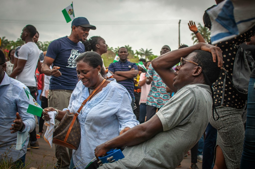 A group of people participating in a lively outdoor event, with a man leaning back and laughing while a woman nearby smiles, both holding small Nigerian flags, creating a joyful and celebratory atmosphere.