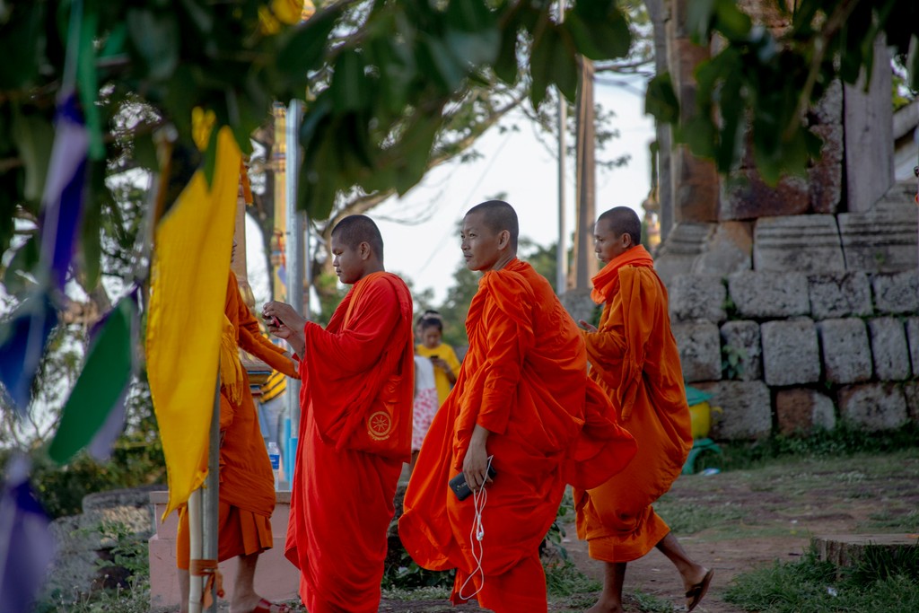 Buddhist monks dressed in vibrant orange robes walk through a temple complex, engaging in their daily rituals amid a serene, nature-filled setting. The scene captures the spiritual devotion and cultural heritage of the monastic life.