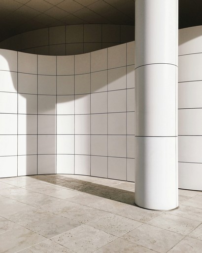 A minimalist interior space featuring a white column and tiled walls with soft shadows on the floor.