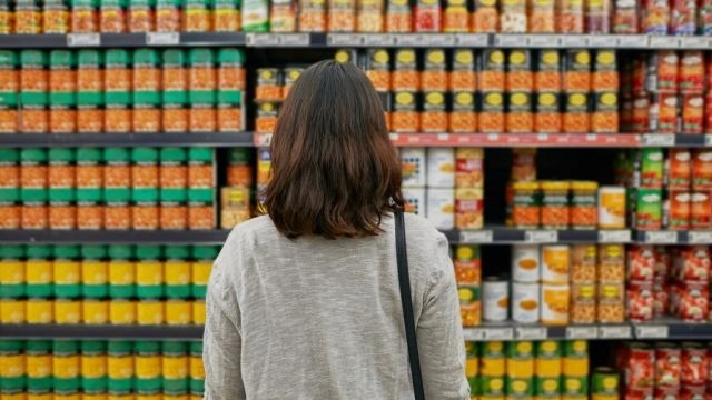 Mulher de costas observando prateleiras cheias de latas e potes organizados em ambiente de mercado.