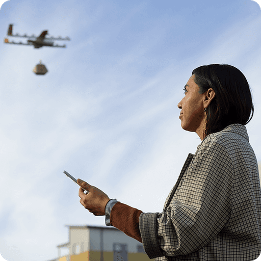 A person standing outdoors operates a smartphone while observing a delivery drone carrying a package, set against a clear blue sky.