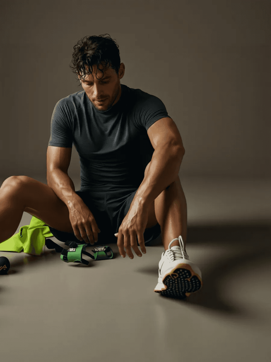 Man sitting on the floor after a workout, wearing a dark athletic outfit with gym equipment beside him.