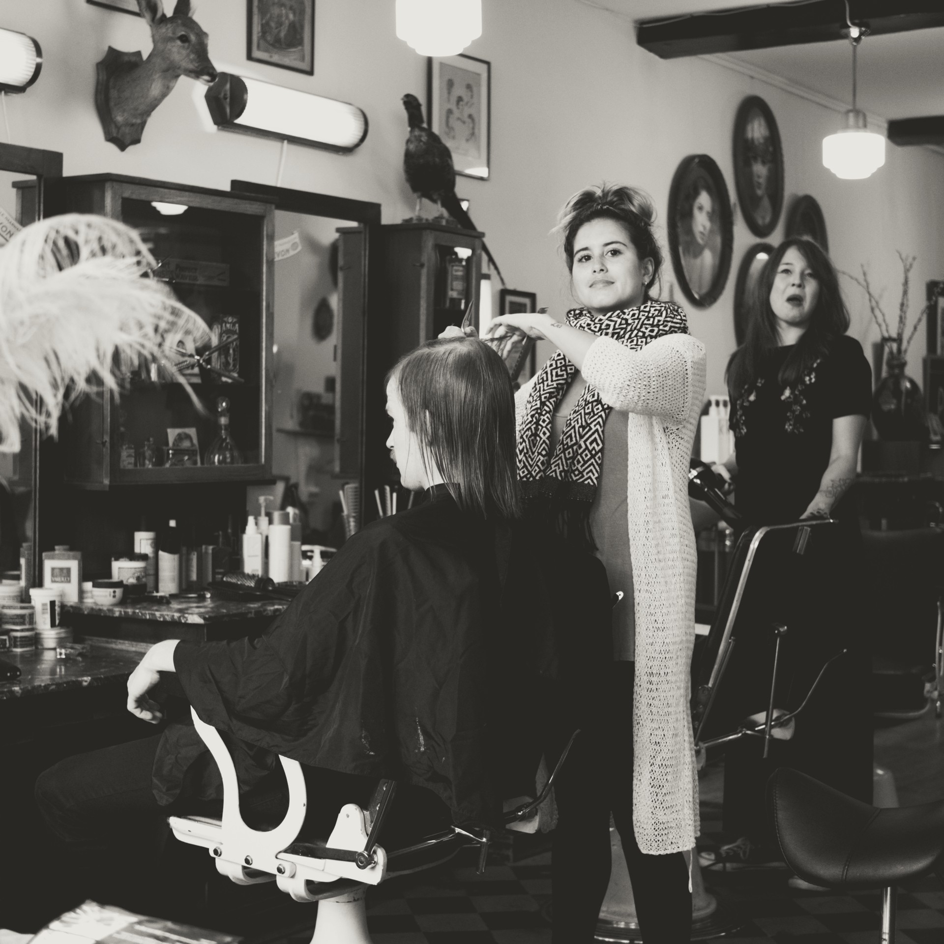 Black-and-white hair salon image showing a stylist cutting a client’s hair in a professional salon interior with mirrors, shelves of hair products, framed decor, and creative styling tools — ideal for content about haircuts, hairstyling services, salon interiors, beauty salons, and professional haircare.
