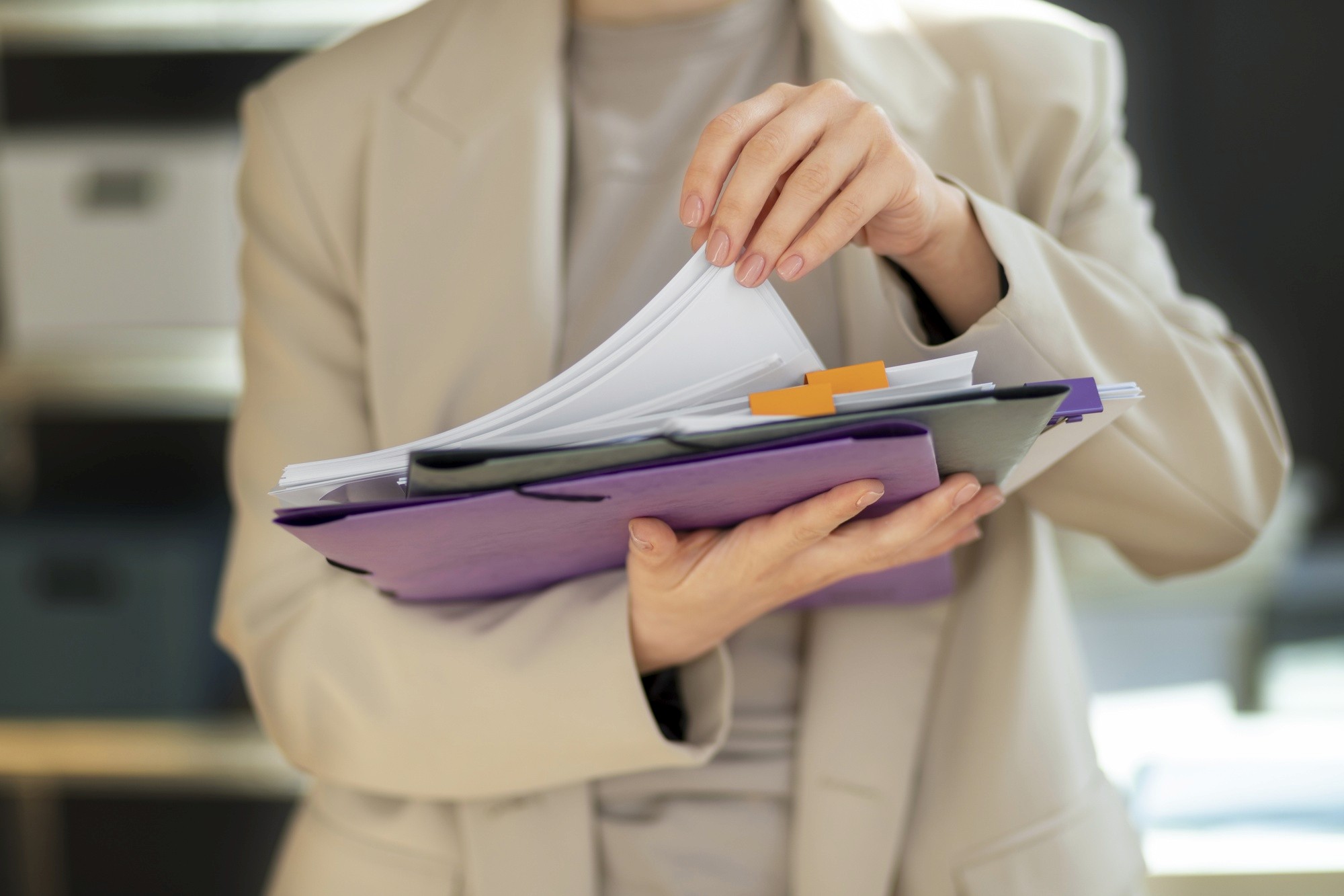 A person in a beige blazer organizing documents and folders, holding papers with colorful tabs