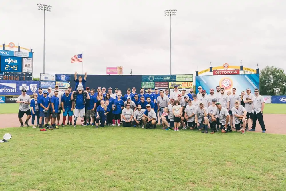 Large group photo of Bernardon (Core States Group) team members on a baseball field, posed in front of stadium advertising boards. Team members are wearing blue and grey uniforms, standing on the outfield grass under overcast skies, with stadium lights and an American flag visible in the background.