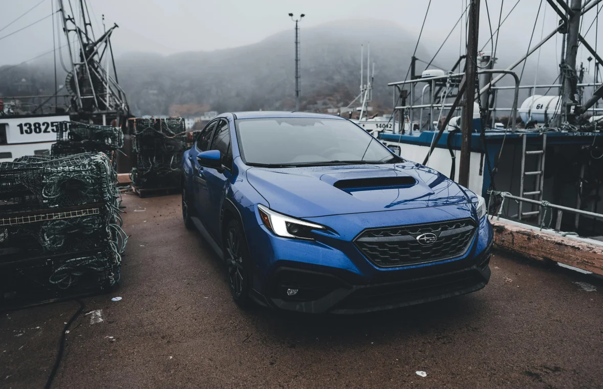 Une Subaru WRX bleue stationnée dans un port enveloppé de brouillard, entourée de casiers de pêche et de bateaux amarrés. L’ambiance froide et humide renforce le caractère robuste et sportif de la berline, prête à affronter n’importe quelle route.