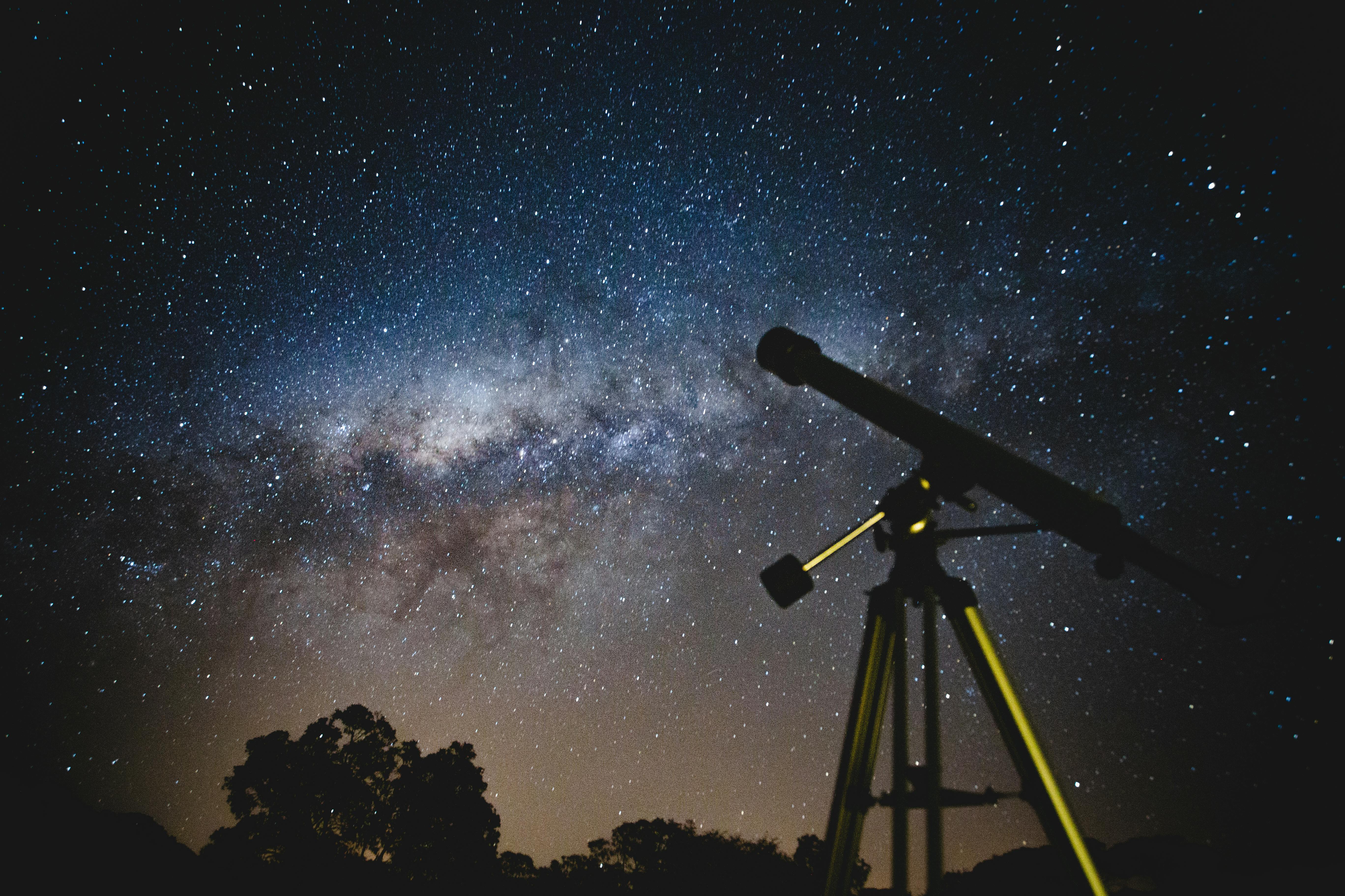 Silhouette of telescope looking up at a starry sky with a nebula in the background