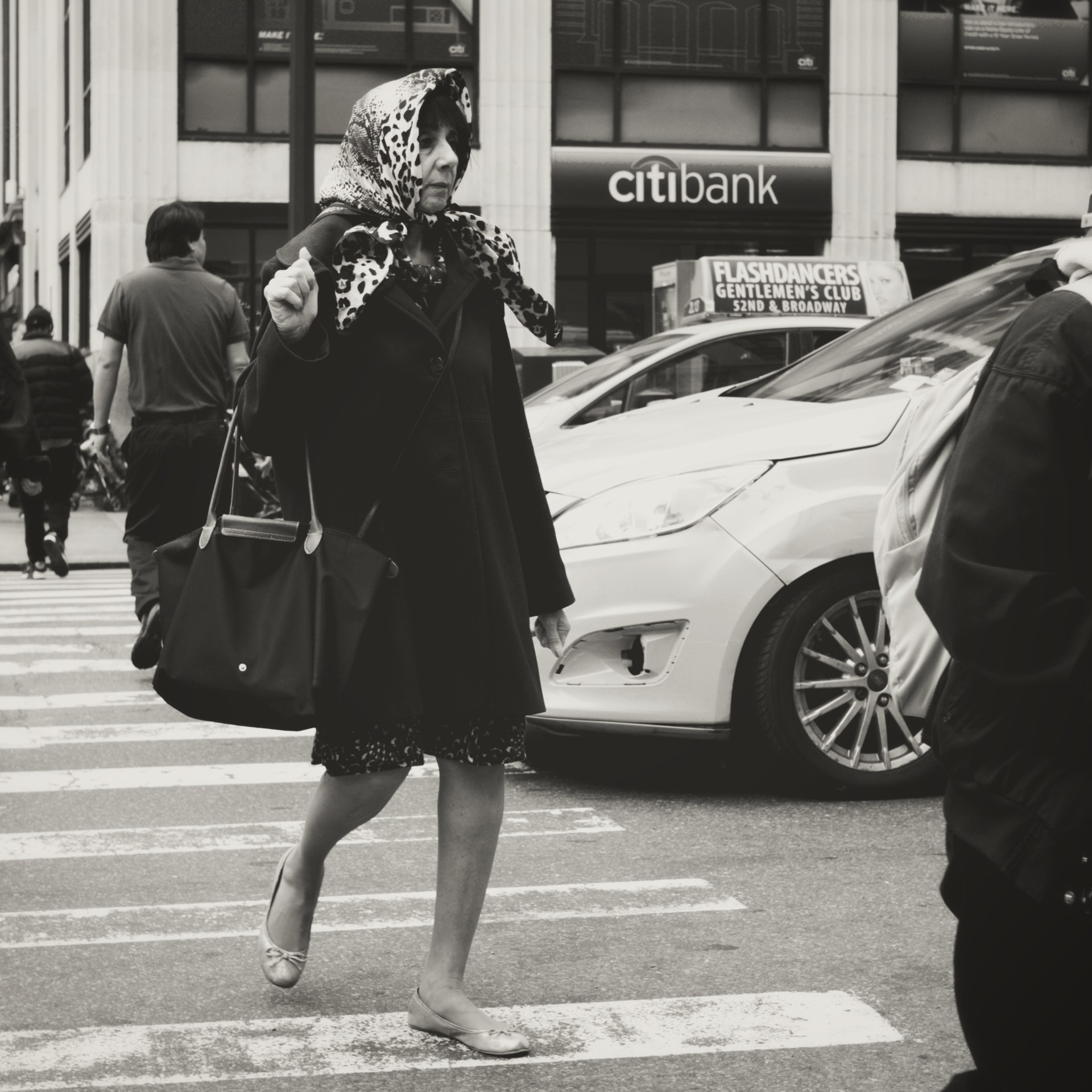 Black-and-white candid image of an older woman in a headscarf and heavy coat crossing a busy urban intersection, capturing a moment of everyday movement against a backdrop of city signage and traffic.