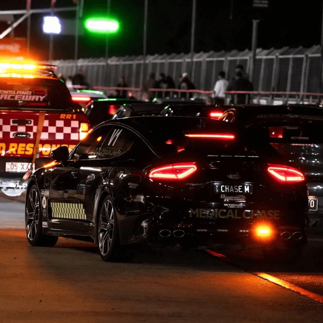 A black medical chase car with glowing red taillights follows a red rapid response vehicle at night, highlighting.