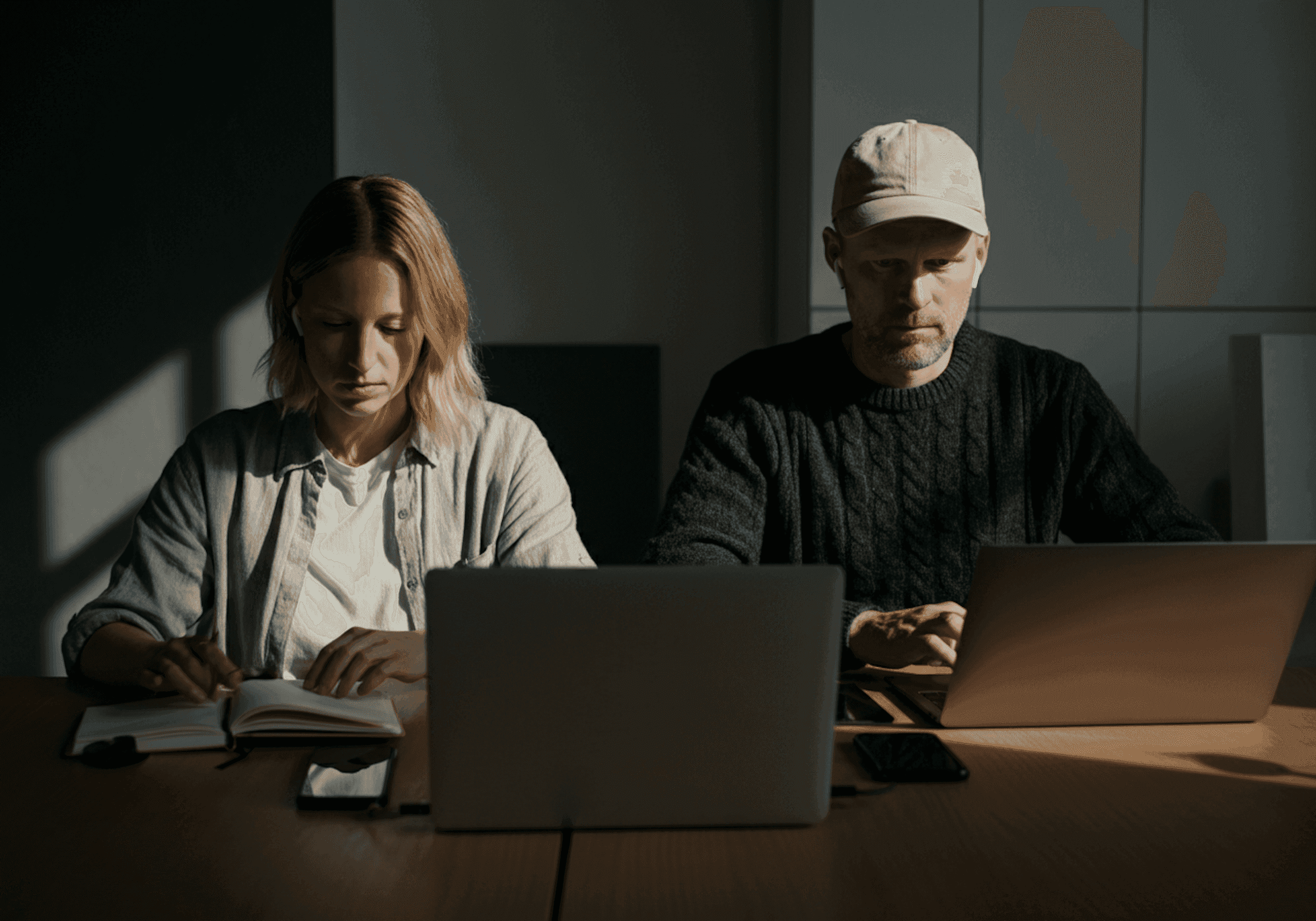 Two people working at a table with laptops, one writing in a notebook. A cozy workspace with natural lighting and a focused atmosphere.