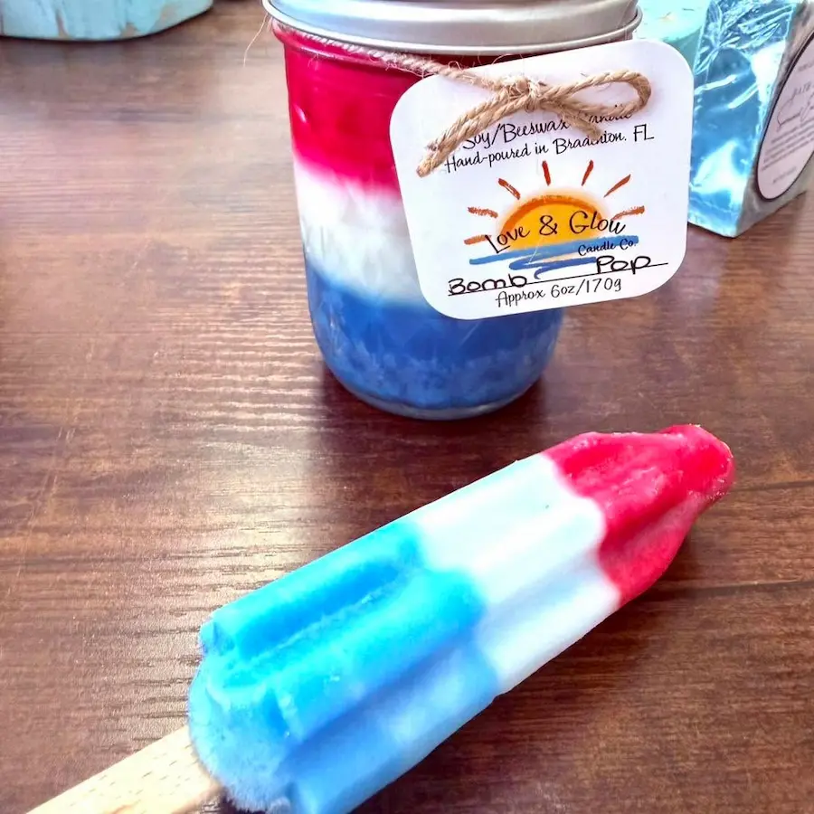 A vibrant red, white, and blue handcrafted popsicle is displayed beside a matching candle jar, both labeled as made in Bradenton, FL, on a wooden surface.