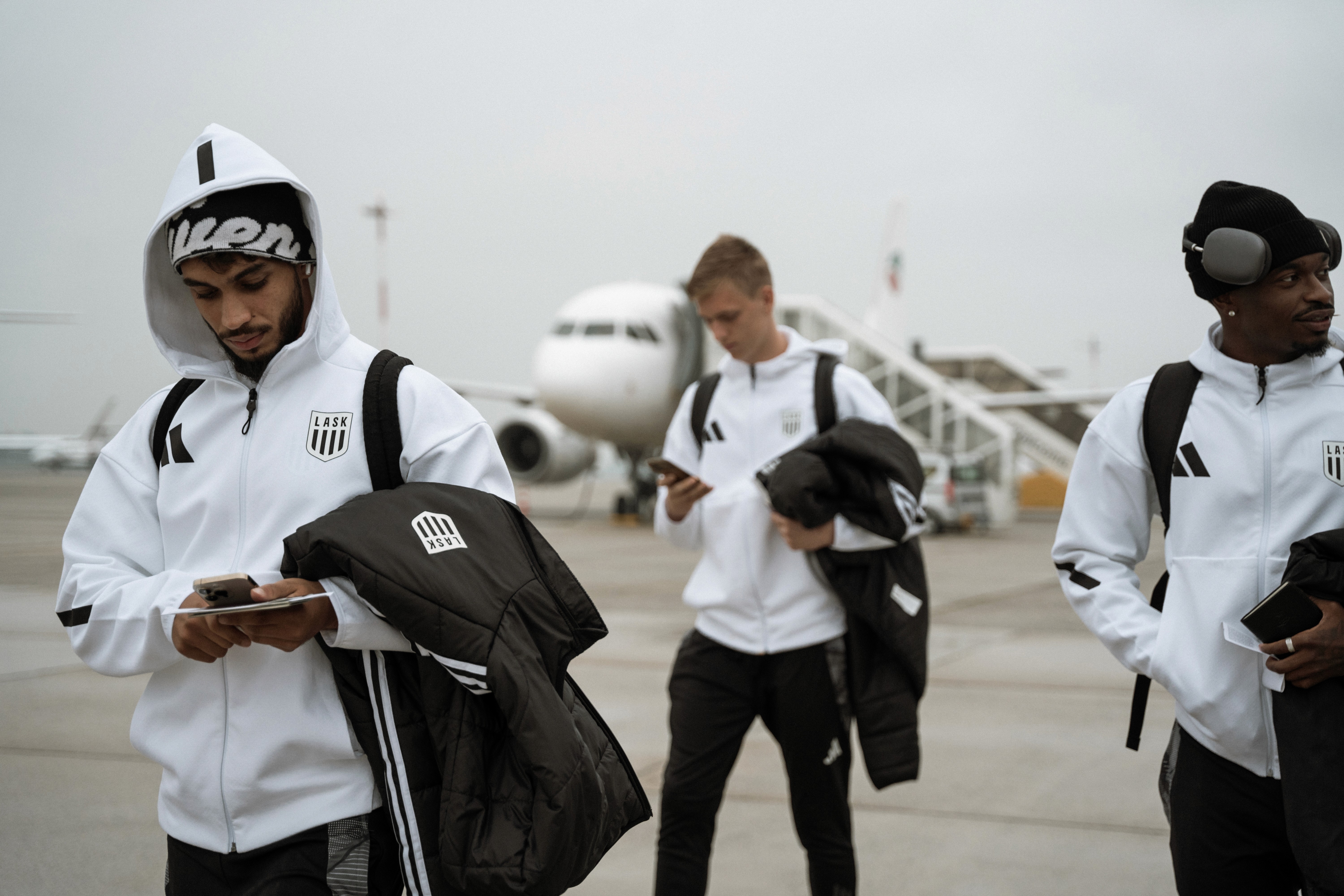 Fußballspieler in weißen Jacken vor einem Flugzeug. Sie tragen Rucksäcke und sind anscheinend auf dem Weg zu einem Spiel.