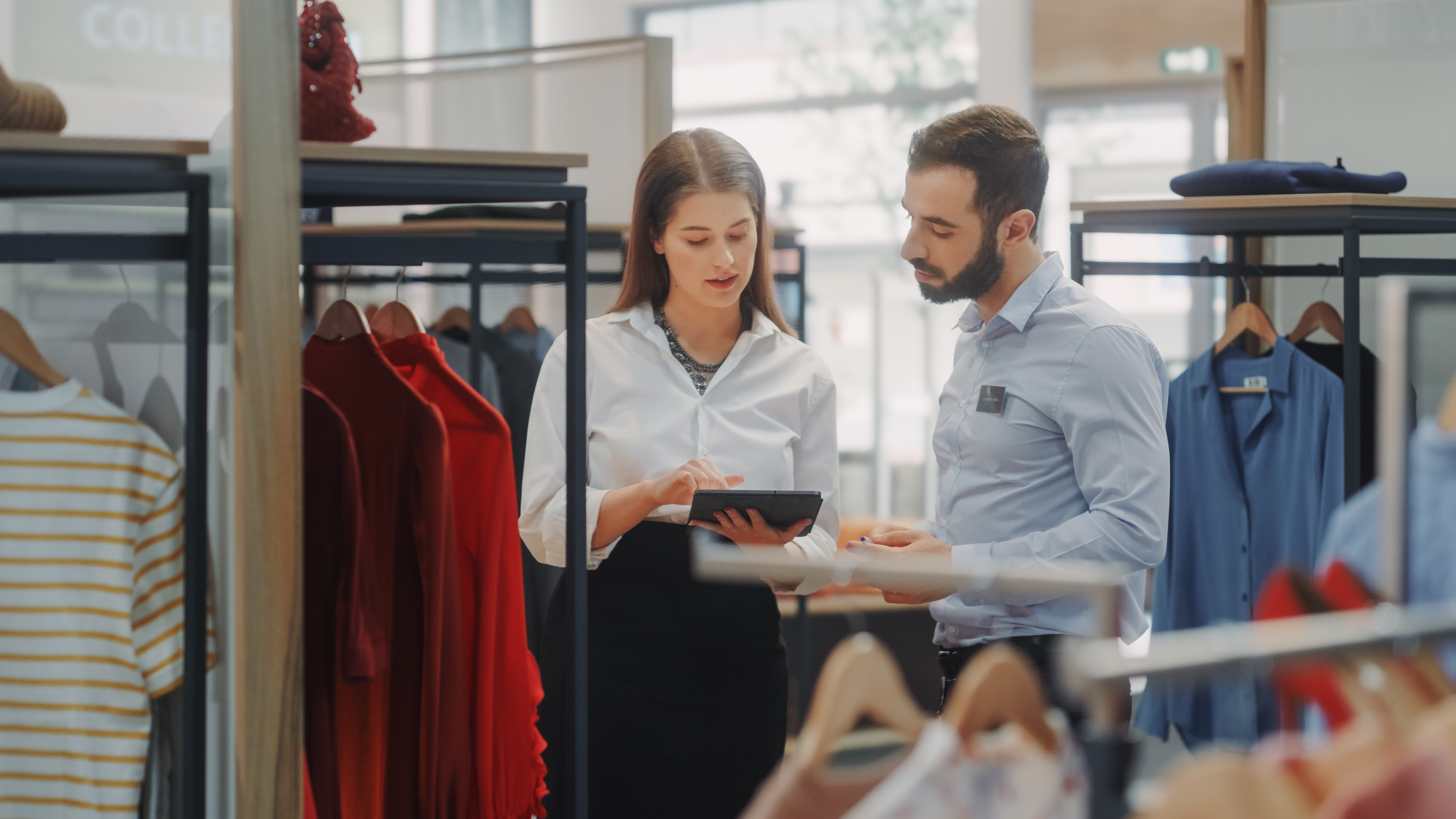 A retail store setting with two employees standing near racks of clothing. One is a woman in a white blouse and black skirt, holding a tablet, while the other is a man in a light blue shirt. They appear to be engaged in a professional discussion about the store's operations.