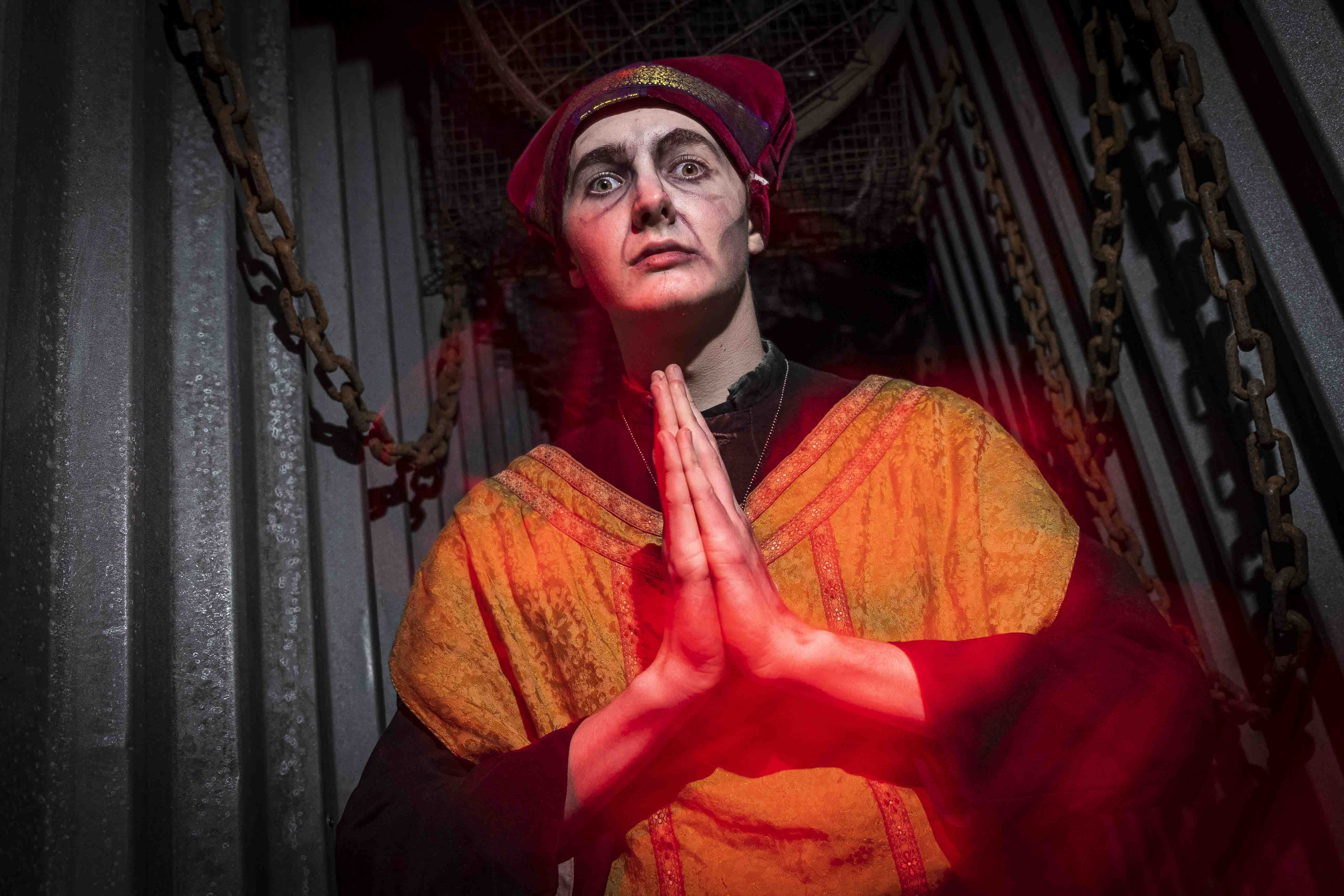 A person wearing theatrical makeup and an elaborate costume, featuring a red hat and a gold-trimmed robe, stands in a dramatic pose with hands clasped, surrounded by chains in a dimly lit industrial setting, illuminated by a mysterious red light.