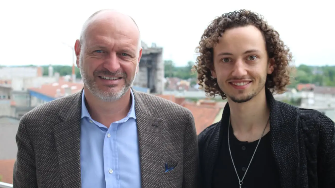 Christian Reindl, dressed in a grey-brown blazer and light blue shirt, stands next to Thiago Calderaro, who is wearing a dark sweater. The two are smiling in front of an urban background with rooftops and a partially visible structure. The image represents their collaboration on CoachingArea, where they share strategies to modernise amateur sports clubs. In the podcast episode they discuss solutions such as professionalising club management, leveraging digital innovation, and developing partnerships to enhance the sustainability of local sports organisations.