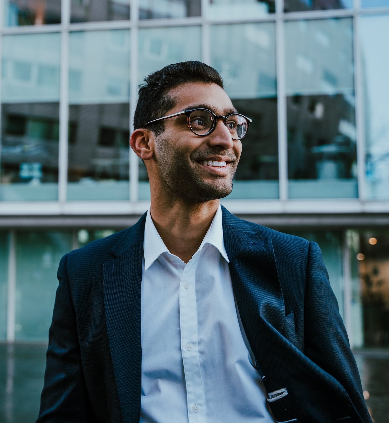 A smiling man wearing glasses, a white shirt, and a navy blazer standing outdoors in front of modern glass buildings.