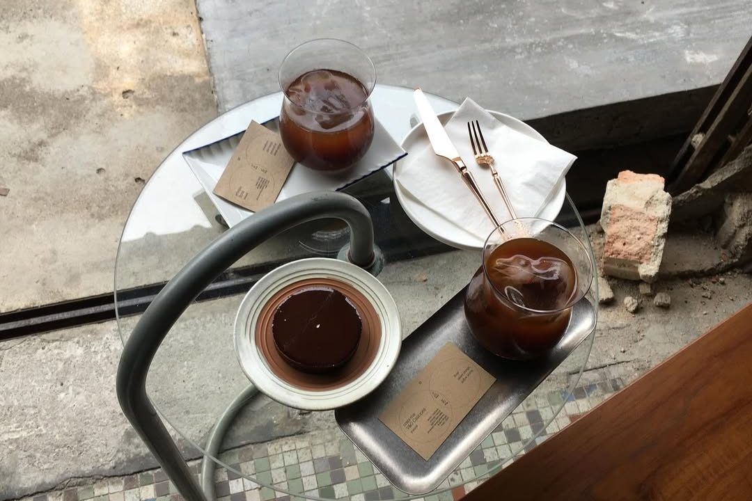 A top-down view of a small cafe table featuring two glasses of iced coffee, a plate with dessert, a white plate, cutlery, and a menu card on a glass tabletop.