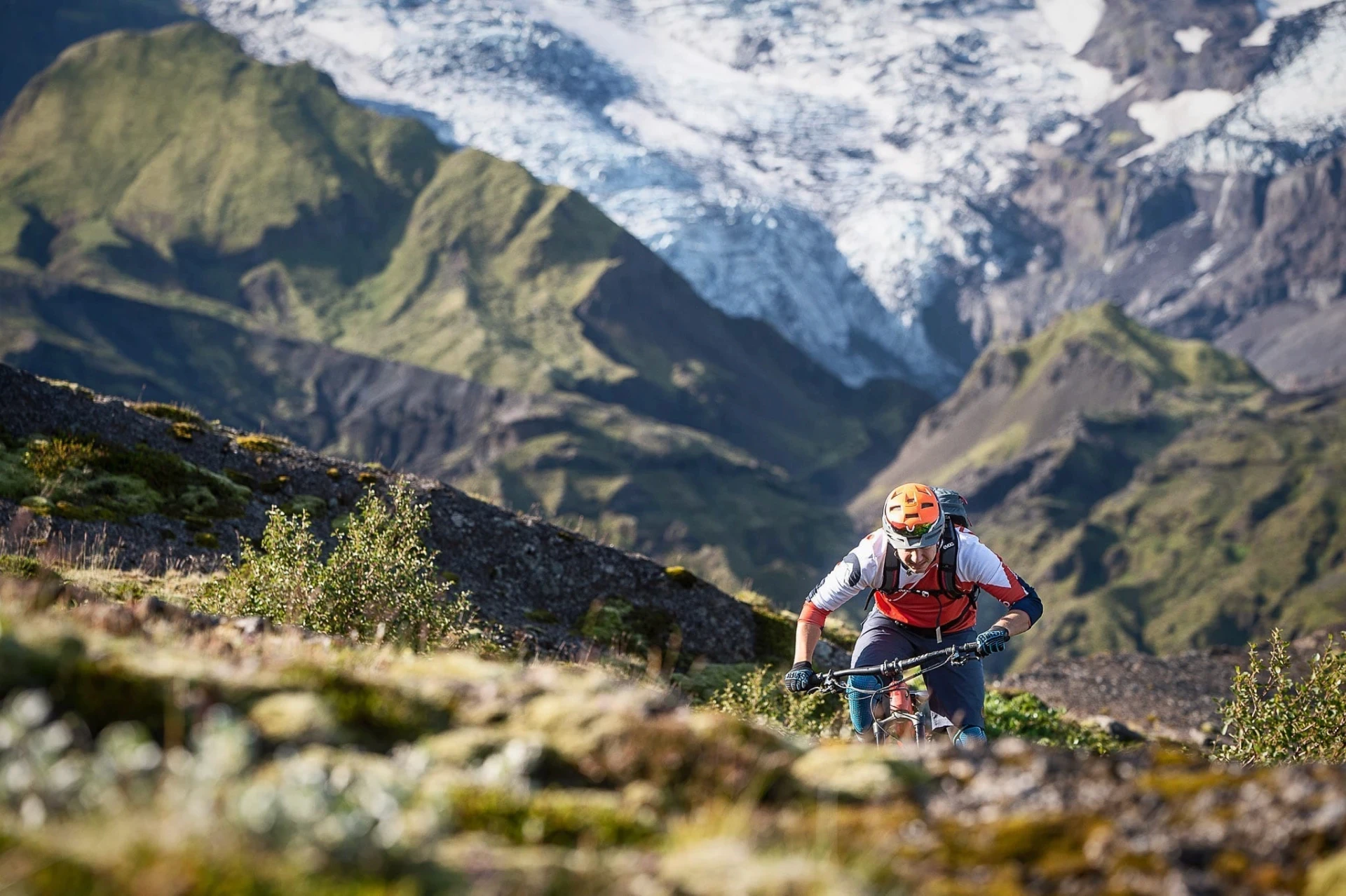 Mountain biker climbing a trail with green mountains and a glacier behind.