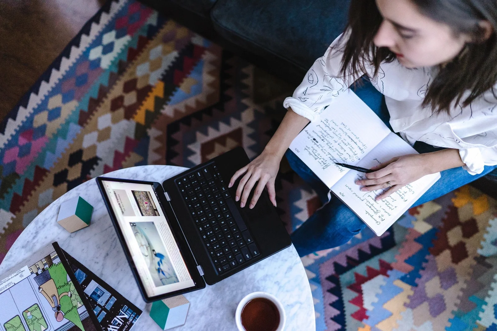 A woman sits on a colorful patterned rug, working on a laptop with a notebook in hand, surrounded by a cozy coffee table topped with a cup of tea, creating a productive work-from-home environment.