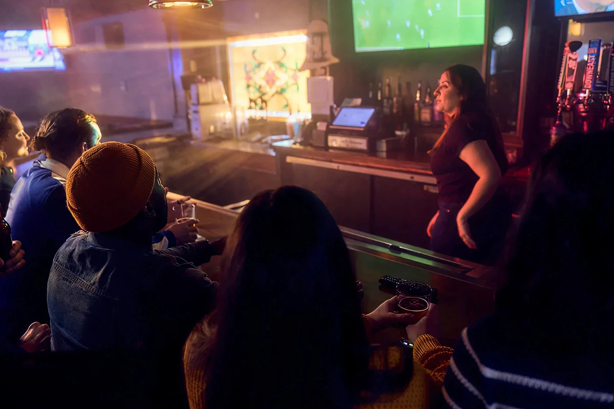 Group of friends and bartender watching live football match on big screen in atmospheric sports bar, warm moody lighting with drinks in hand and pool table in foreground"