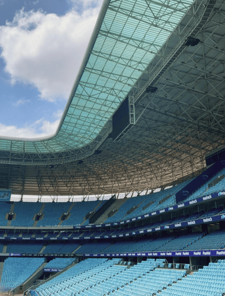 Vista interna ddo estádio do Grêmio vazio, mostrando as arquibancadas azuis e parte da cobertura contra um céu com nuvens.