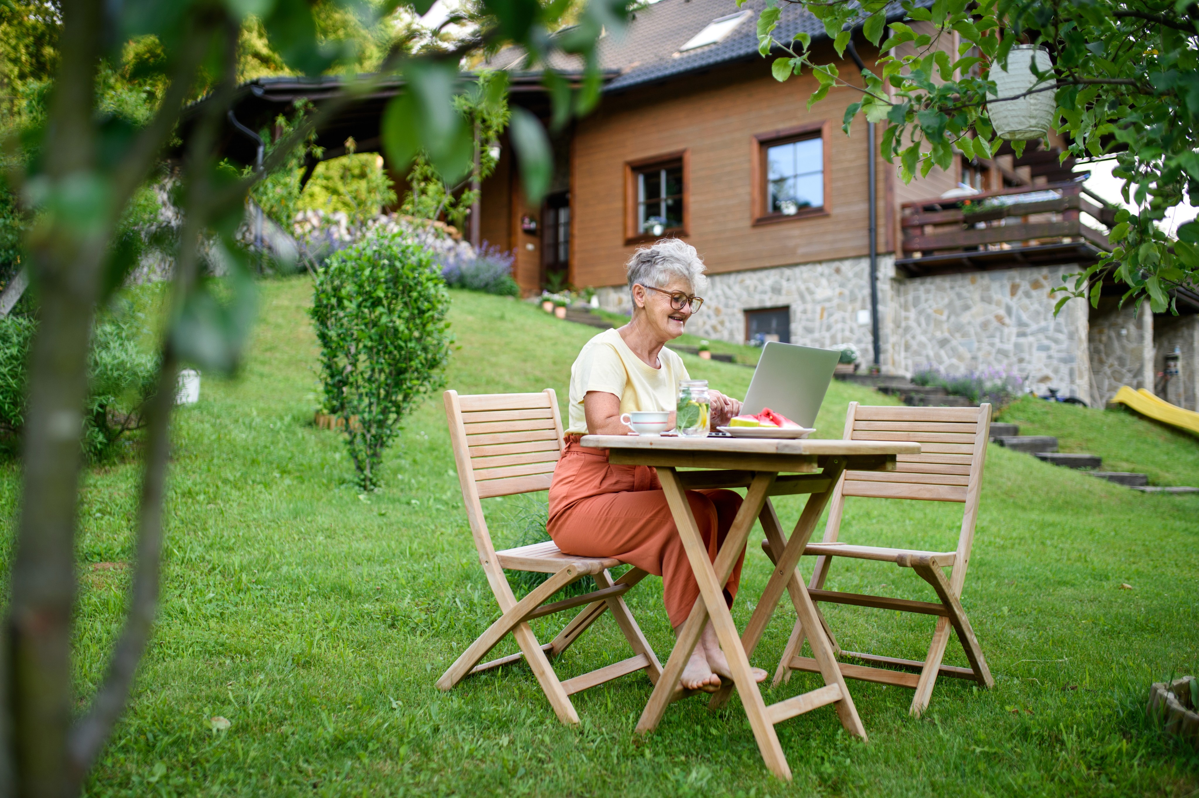 Mulher idosa sentada na frente de um notebook sobre uma mesa em um jardim de grama e árvores