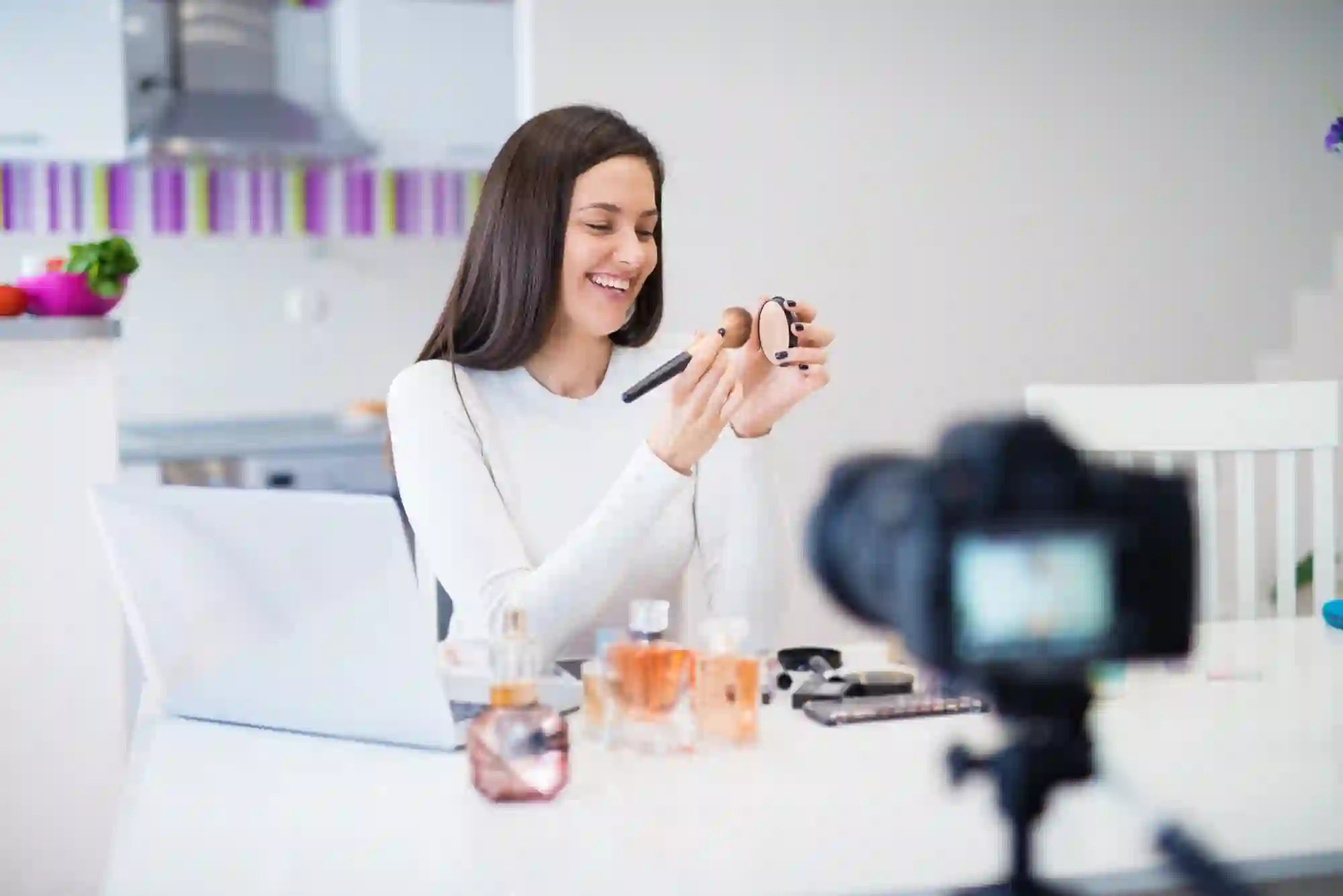 A smiling beauty influencer is filming a tutorial in her kitchen, holding a makeup brush and compact in front of a camera.
