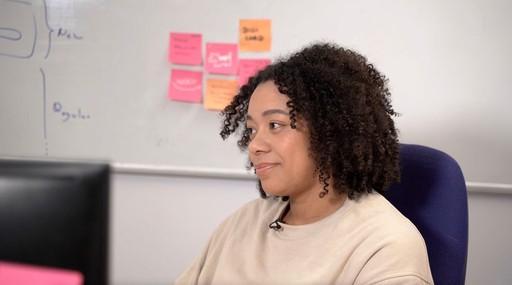 A person with curly hair sits at a desk, looking thoughtfully to the side in a modern office environment.