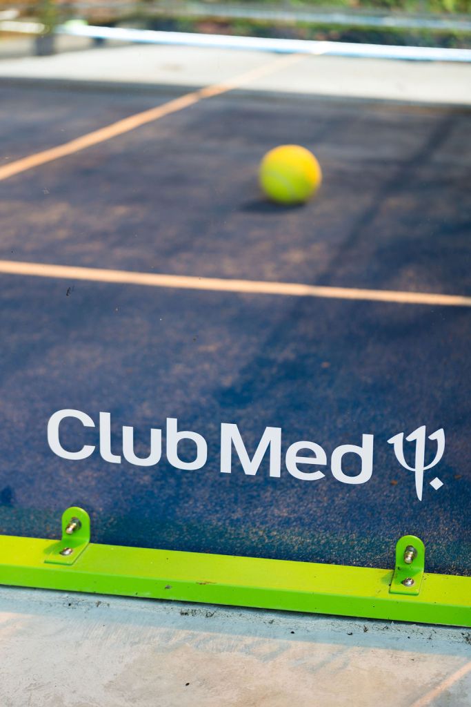 A close-up view of a tennis court featuring the Club Med logo, with a yellow tennis ball in motion on the court's surface.