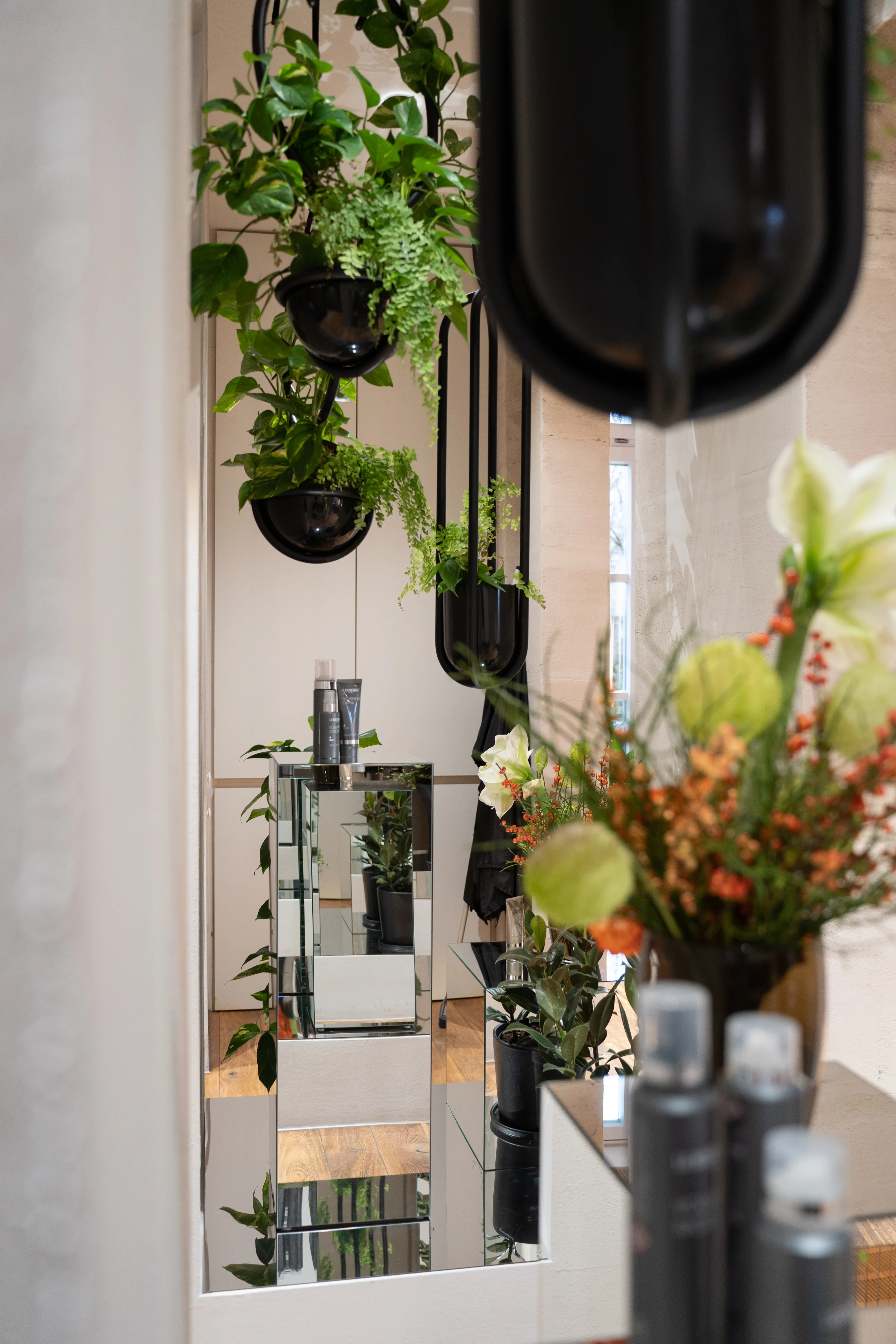 Hanging black planters with green plants above a mirrored product display cube at EMPL FRISEURE, with flowers in the foreground.