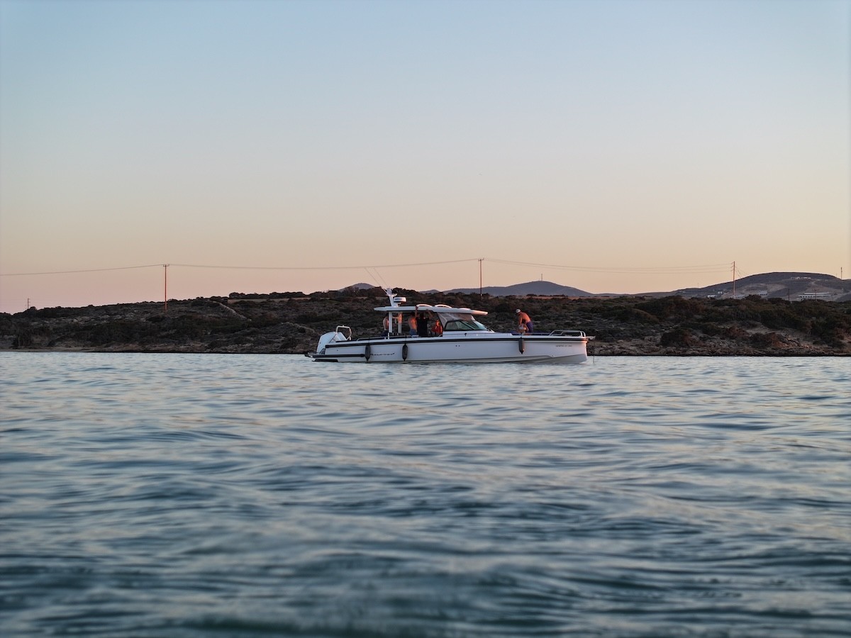 White Axopar 37 motor yacht with passengers aboard during golden hour sunset, anchored in calm waters with rocky coastline silhouetted in background.