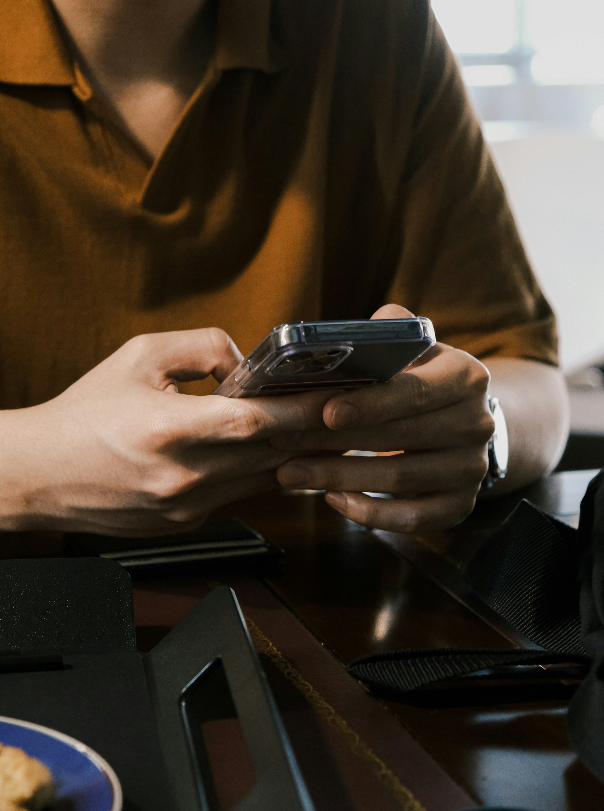 A person's hands are using a clear-cased smartphone while holding it close to a table in a brightly lit environment.
