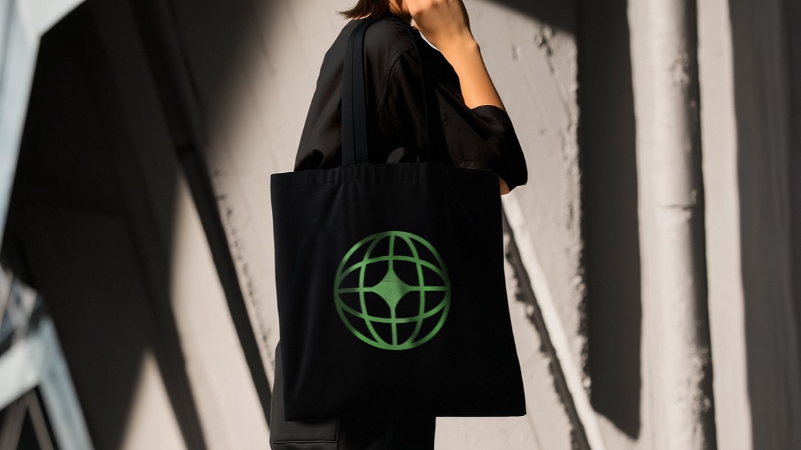 Person wearing black clothing carrying a black tote bag with a green globe-like geometric symbol, standing against a concrete wall in sunlight and shadow.