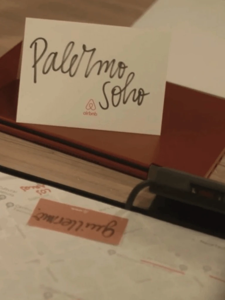 Tarjeta de Airbnb con “Palermo Soho” escrito a mano, apoyada en bandeja roja sobre escritorio.